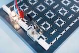Auf der Oberfläche eines Experimentierboards sind elektrische Bauteile wie Glühbirnen und Schalter sichtbar, die mit bunten Kabeln verbunden sind. Das Board verfügt über nummerierte Anschlusslöcher, um verschiedene Schaltungen zu erstellen und zu testen.