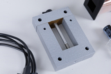 Ein graues Metallgehäuse mit einer offenen Aussparung und einer senkrechten Metallstange ist im Fokus. Daneben sind einige Kabel und ein elektronisches Bauteil zu sehen, die für Experimente im Bildungsbereich genutzt werden könnten.