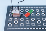 Auf der Platine sind eine Spule und ein rotierendes Element mit grünen und roten Flügeln zu sehen, die durch einen elektrischen Antrieb in Bewegung versetzt werden. Das Design der Platine mit vielen Löchern ermöglicht eine flexible Anordnung der Komponenten für Experimente.