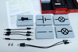 Auf dem Tisch liegen mehrere elektronische Bauteile in grauen Gehäusen, die verschiedene Schaltsymbole anzeigen. Zudem sind einige Kabel mit Steckverbindungen und zwei Glühbirnen vorhanden, sowie ein bereits geöffnetes Handbuch mit Schaltplänen zur Erklärung.