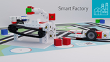 In einer modernen Modellfabrik sind verschiedene Bauelemente und Miniaturroboter gestaltet, die eine Produktionslinie darstellen. Auf einem farbenfrohen Spielplan sind Würfel und ein Roboterarm zu sehen, die zusammen eine smarte Fertigungsumgebung simulieren.