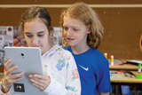 Zwei Kinder stehen in einem Klassenzimmer und schauen gemeinsam auf ein Tablet. Die Umgebung ist freundlich gestaltet, mit Bastelmaterialien und farbigen Gegenständen auf einem Tisch im Hintergrund.