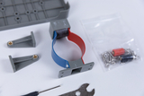 Auf der Oberfläche sind verschiedene Komponenten eines Experimentierkits angeordnet. Zu sehen sind unter anderem eine Halterung mit rot-blauer Umrandung, Schrauben, eine Werkzeugklemme und weitere Bauteile in einer Verpackung.