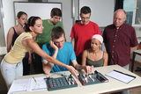 Eine Gruppe von fünf Personen arbeitet gemeinsam an einem Experimentier-Set auf einem Tisch. Sie scheinen sich auf die Bedienung des Geräts zu konzentrieren, während ein Lehrer oder Betreuer das Geschehen beobachtet.