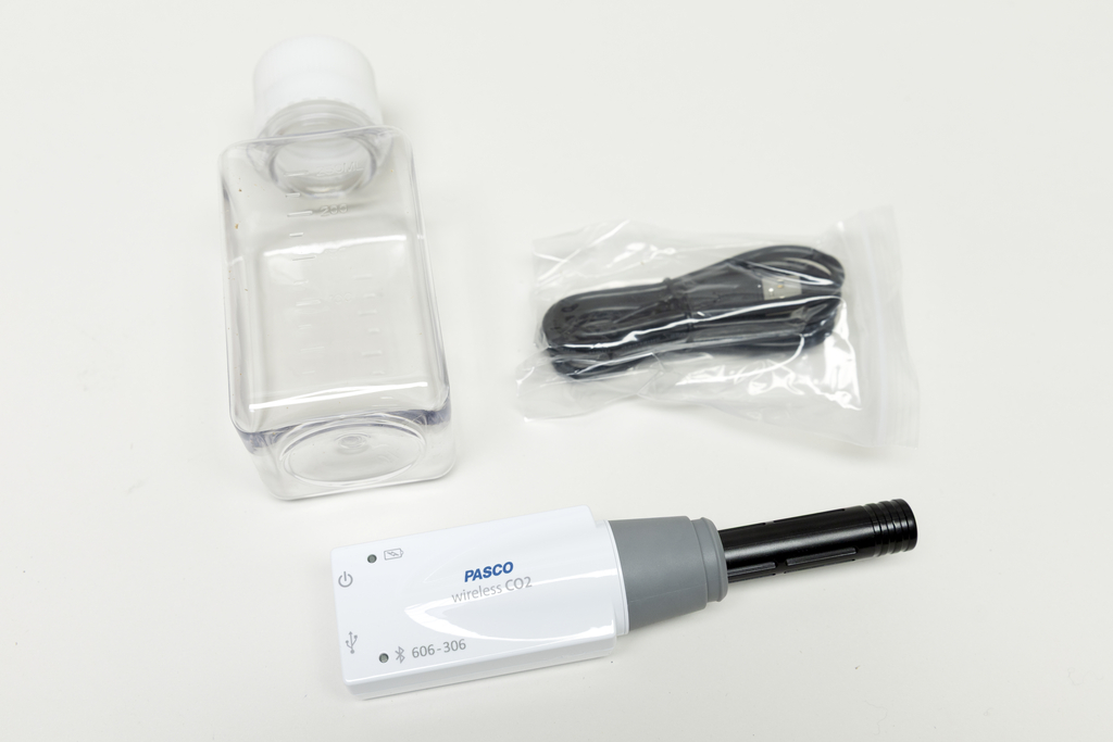 Ein drahtloser CO2-Sensor von PASCO liegt neben einer leeren Kunststoffflasche. Ein verpacktes USB-Ladekabel ergänzt das Set für wissenschaftliche Experimente.