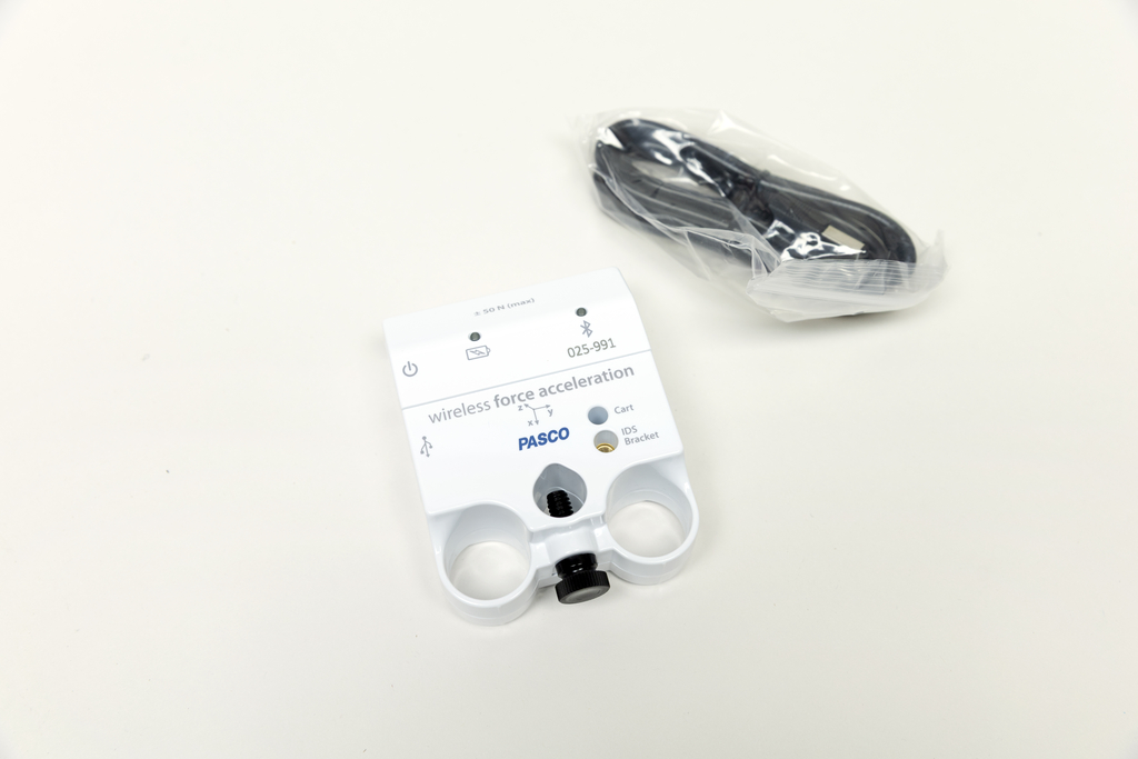 Ein weißer PASCO-Sensor zur Messung von Kraft und Beschleunigung liegt auf einer hellen Fläche. Daneben befindet sich ein in Plastik verpacktes schwarzes Verbindungskabel.