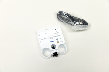 Ein weißer PASCO-Sensor zur Messung von Kraft und Beschleunigung liegt auf einer hellen Fläche. Daneben befindet sich ein in Plastik verpacktes schwarzes Verbindungskabel.