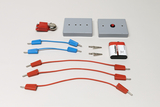 Ein Set aus elektrischen Bauteilen umfasst eine Batterie, eine Glocke und zwei graue Module. Ergänzt wird das Sortiment durch rote und blaue Verbindungskabel sowie zwei Krokodilklemmen.