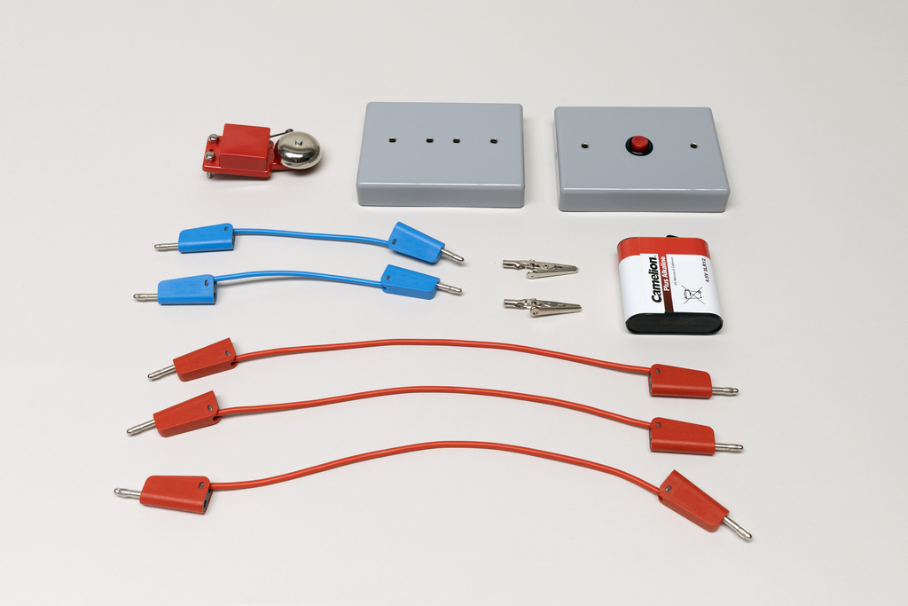 Ein Set aus elektrischen Bauteilen umfasst eine Batterie, eine Glocke und zwei graue Module. Ergänzt wird das Sortiment durch rote und blaue Verbindungskabel sowie zwei Krokodilklemmen.
