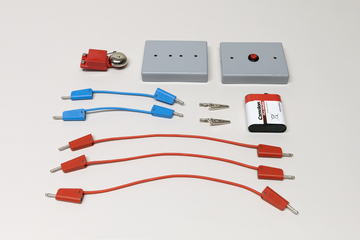 Ein Set aus elektrischen Bauteilen umfasst eine Batterie, eine Glocke und zwei graue Module. Ergänzt wird das Sortiment durch rote und blaue Verbindungskabel sowie zwei Krokodilklemmen.
