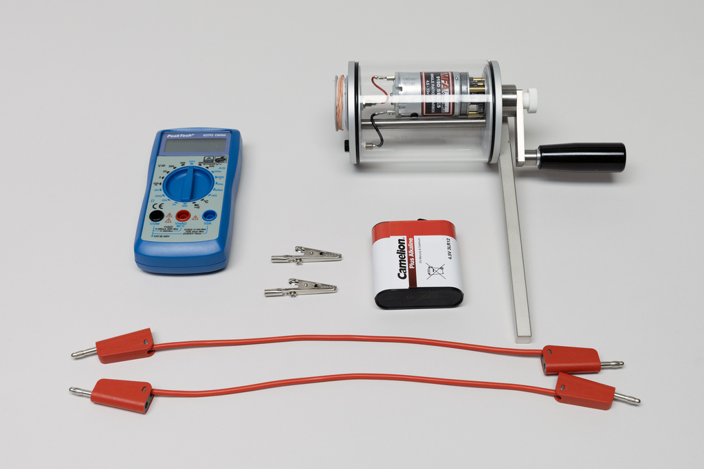 Ein blaues Digitalmultimeter, eine Handkurbel-Dynamo-Einheit und eine Batterie liegen auf einer hellen Fläche. Ergänzt wird das Set durch zwei Krokodilklemmen sowie zwei rote Verbindungskabel.