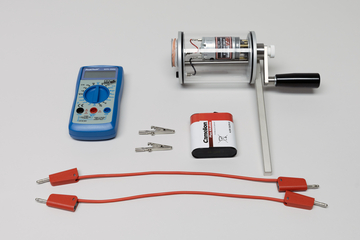 Ein blaues Digitalmultimeter, eine Handkurbel-Dynamo-Einheit und eine Batterie liegen auf einer hellen Fläche. Ergänzt wird das Set durch zwei Krokodilklemmen sowie zwei rote Verbindungskabel.