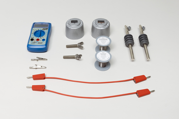 Ein blaues Multimeter liegt neben verschiedenen metallischen Bauteilen, Spulen und zwei roten Verbindungskabeln. Das Set umfasst zudem Krokodilklemmen sowie isolierte Kontaktstifte für physikalische Experimente.
