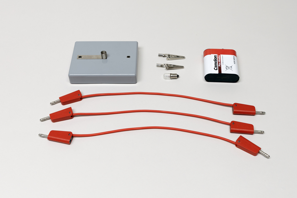Ein grauer Batteriekasten, drei rote Anschlusskabel und eine Batterie liegen auf einer weißen Fläche. Ergänzt wird das Set durch zwei Krokodilklemmen und eine kleine Glühbirne.