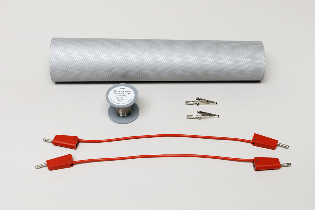 Eine Rolle Konstantandrat, zwei Krokodilklemmen und zwei rote Verbindungskabel liegen auf hellem Hintergrund. Diese Materialien dienen als Versuchsset für physikalische Experimente im Bildungsbereich.