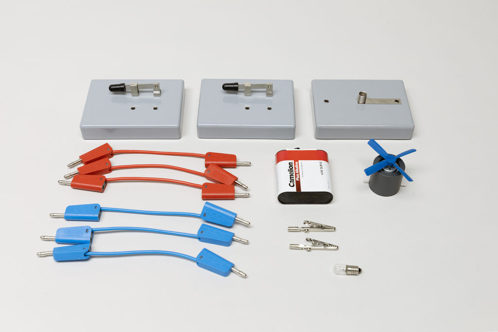 Ein physikalisches Experimentierset enthält drei graue Basismodule, rote und blaue Verbindungskabel sowie eine Batterie. Ergänzt wird das Set durch zwei Krokodilklemmen, eine kleine Glühbirne und einen blauen Ventilator.