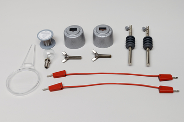 Ein Physik-Experimentierset umfasst zwei metallene Halterungen, Schrauben, Klemmen und zwei rote Verbindungskabel. Ergänzt wird das Set durch eine Glühbirne, eine Spule und eine Lupe.
