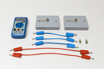 Ein blaues Digitalmultimeter liegt neben zwei grauen Prüfmodulen auf weißem Grund. Ergänzt wird das Set durch diverse rote und blaue Verbindungskabel sowie kleine Metallstifte.