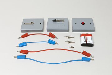 Ein physikalisches Experimentierset umfasst drei graue Module mit Schaltern sowie rote und blaue Anschlusskabel. Ergänzt wird das Set durch Krokodilklemmen und eine Batterie.