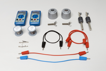 Ein Set physikalischer Experimentiergeräte umfasst zwei Multimeter, verschiedene Verbindungskabel und Klemmen. Ergänzt wird das Ensemble durch metallische Bauteile sowie zwei zylindrische Halterungen mit Beschriftung.
