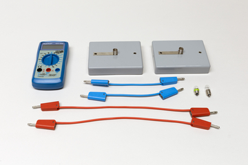Ein blaues Digitalmultimeter liegt neben zwei grauen Modulen und mehreren Anschlusskabeln in Blau und Rot. Ergänzt wird das Set durch zwei kleine Glühbirnen.