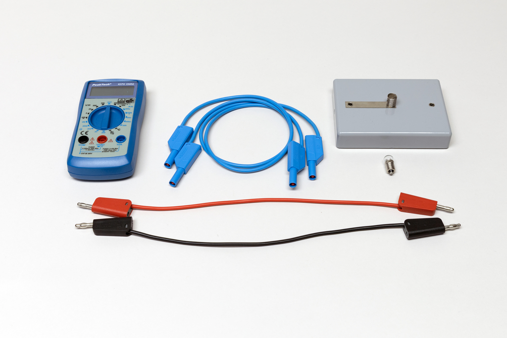 Ein blaues Digitalmultimeter liegt neben verschiedenen Anschlusskabeln in Blau, Rot und Schwarz. Rechts daneben befinden sich ein grauer Messblock sowie eine kleine Glühbirne.