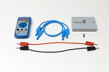 Ein blaues Digitalmultimeter liegt neben verschiedenen Anschlusskabeln in Blau, Rot und Schwarz. Rechts daneben befinden sich ein grauer Messblock sowie eine kleine Glühbirne.