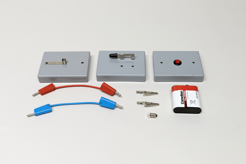 Ein Set aus drei grauen Modulen mit Schaltern und Kontakten liegt auf einer weißen Fläche. Ergänzt wird das Experimentierkit durch rote und blaue Kabel, Krokodilklemmen sowie eine 4,5V-Batterie.
