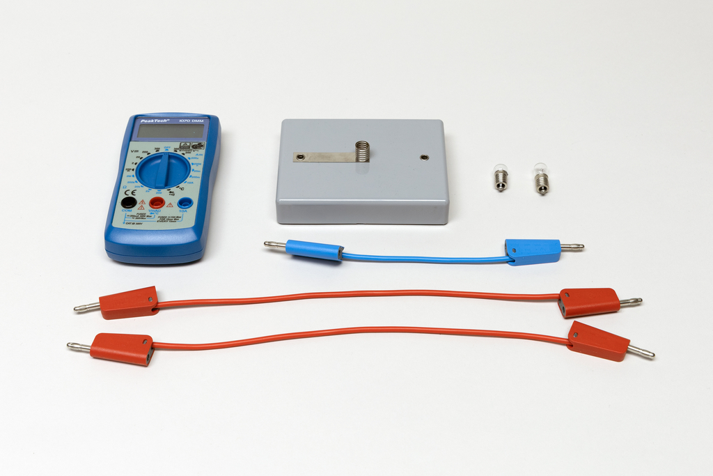 Ein blaues Digitalmultimeter liegt neben einem grauen Gehäuse und zwei kleinen Leuchtmitteln. Drei Verbindungskabel in Blau und Rot ergänzen das Set für elektrische Experimente.