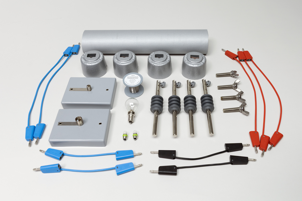 Ein Set aus physikalischen Experimentierutensilien liegt auf hellem Grund. Es umfasst Kabel, Glühbirnen, Metallzylinder, Klemmen und Schalter zur Demonstration elektrischer Leitfähigkeit.