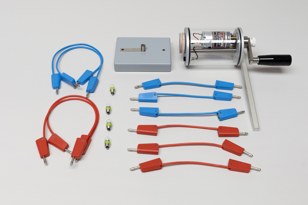 Ein Dynamo mit Kurbel, ein grauer Sockel sowie rote und blaue Anschlusskabel liegen auf weißem Grund. Ergänzt wird das Set durch mehrere kleine Glühbirnen für physikalische Experimente.