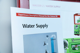 Ein Handbuch für ein Wissenschafts-Kit trägt den Titel „Water Supply“. Darunter ist ein Foto einer Versuchsanordnung mit Wasserhähnen und einem Messzylinder zu sehen.