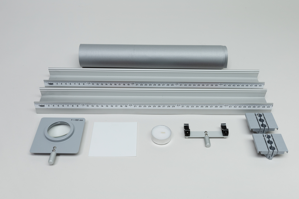Ein Set aus optischen Experimentiergeräten liegt auf einer hellen Fläche. Es umfasst eine optische Bank mit Linealen, eine Linse, einen Spiegel sowie diverse Halterungen und Zubehör.