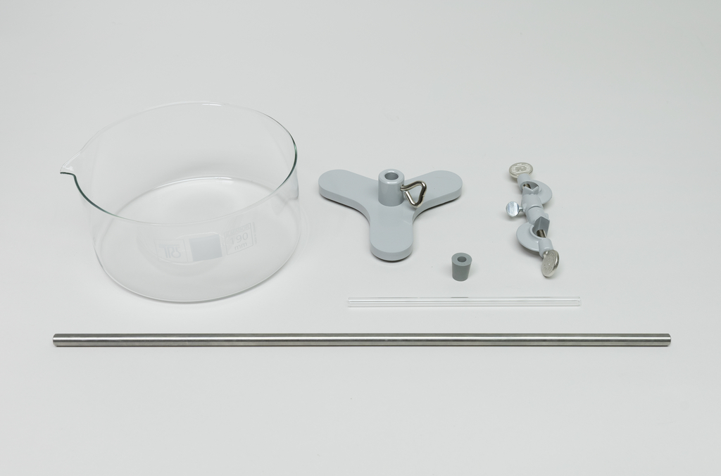 Ein Becherglas aus Glas liegt neben grauen Halterungen und einer kleinen Hülse. Ergänzt wird das Set durch einen Glasstab und eine Metallstange.