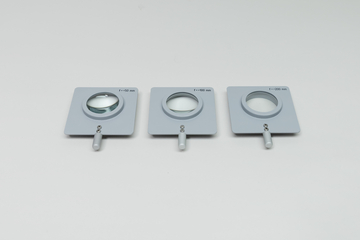 Drei optische Sammellinsen in grauen quadratischen Fassungen liegen nebeneinander. Die Linsen sind mit verschiedenen Brennweiten von 50 mm, 100 mm und 200 mm beschriftet.