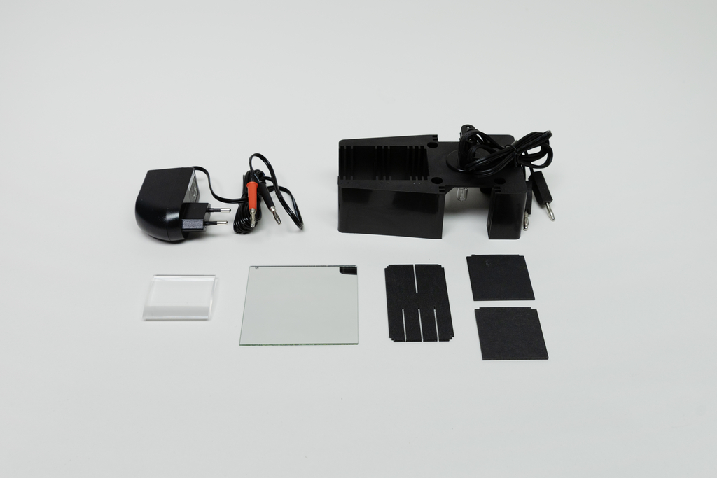 Ein schwarzes Elektrogerät mit Netzteil und Kabeln liegt auf einem hellen Untergrund. Daneben befinden sich diverse Glasplättchen und schwarze Folien für physikalische Experimente.