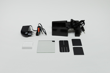 Ein schwarzes Elektrogerät mit Netzteil und Kabeln liegt auf einem hellen Untergrund. Daneben befinden sich diverse Glasplättchen und schwarze Folien für physikalische Experimente.