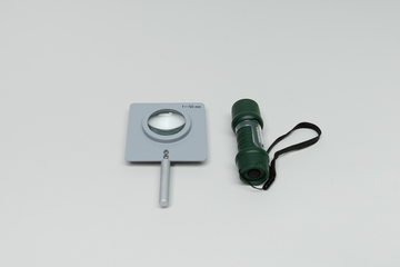 Eine Sammellinse mit einer Brennweite von 50 mm ist in einer grauen Halterung fixiert. Daneben liegt eine kleine, grüne Taschenlampe mit einem schwarzen Trageband.