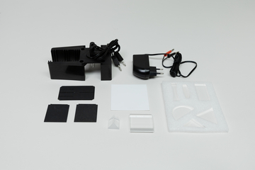 Ein Set aus physikalischen Experimentierutensilien liegt auf einer weißen Fläche. Es umfasst eine Stromquelle mit Adapter sowie diverse Lichtfilter- und Prismenelemente.