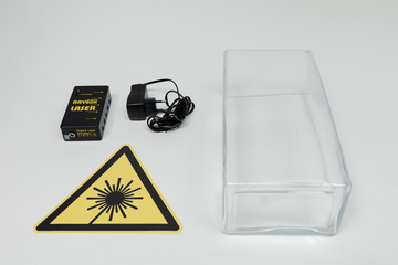 Ein Lasergerät der Marke Raybox liegt neben einem passenden Netzteil und einer transparenten Kunststoffwanne. Ein gelbes Warnschild weist auf die Laserstrahlung hin.