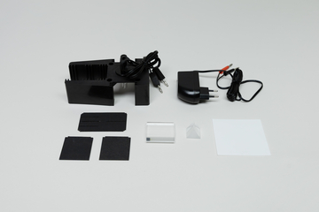 Ein schwarzer Halter mit Netzteil und Kabeln liegt neben verschiedenen Zubehörteilen. Dazu gehören dunkle Quadrate, ein transparenter Glaswürfel und ein weißes Blatt Papier.